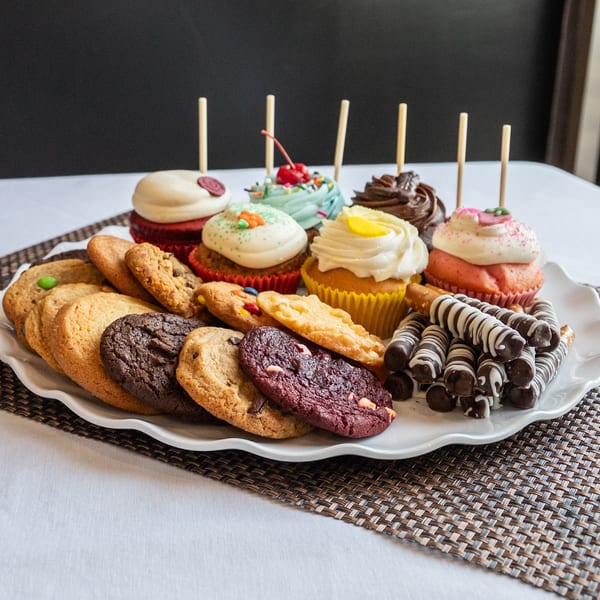 A white platter filled with assorted cupcakes, cookies, and chocolate-dipped pretzel rods on a woven placemat.