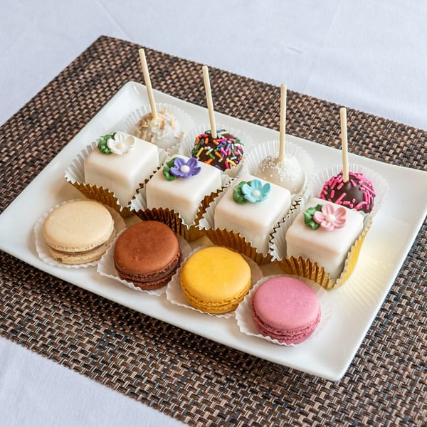 A white rectangular plate holds an assortment of colorful macarons, petit fours, and cake pops.
