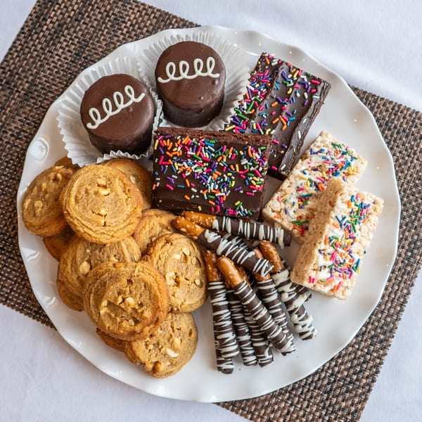 A white platter filled with an assortment of desserts, including cookies, brownies, and chocolate-covered pretzels.