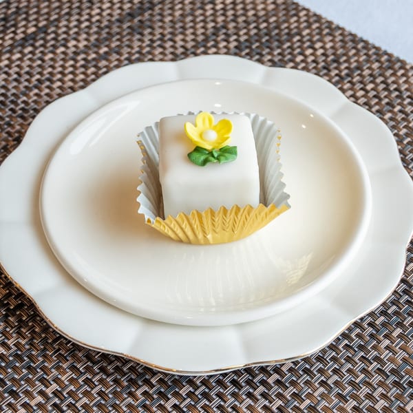 A white petit four with a yellow flower decoration sits in a gold cup on a white plate.