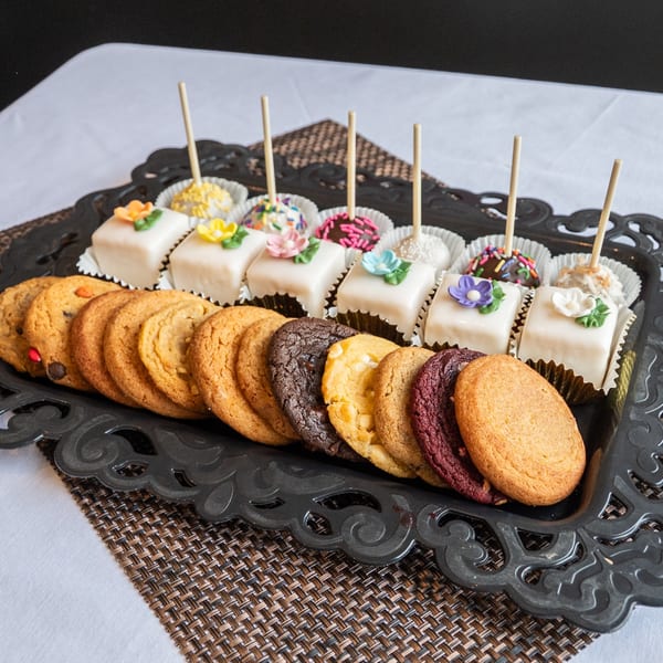 A black ornate tray holds a variety of cookies, petit fours, and cake pops on a white tablecloth.
