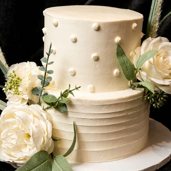 Two-tiered white cake with white roses, green leaves, and textured frosting on a dark background.