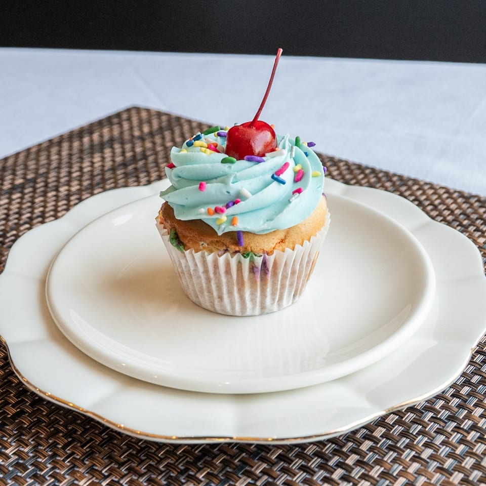 A vanilla cupcake with light blue frosting, colorful sprinkles, and a cherry on top, on white plates.
