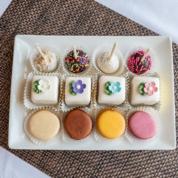 A white rectangular plate holds four cake pops, four petit fours, and four macarons.