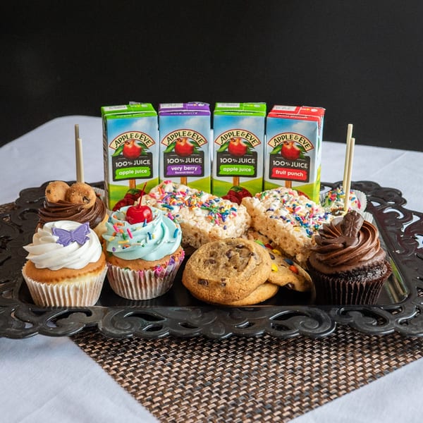 A black tray holds cupcakes, cookies, rice crispy treats, and Apple & Eve juice boxes.